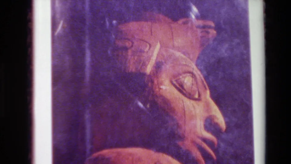 1980 Big Sur 8mm frame holding on a printed or displayed brown carved face, with white page edge and dark purple background visible.