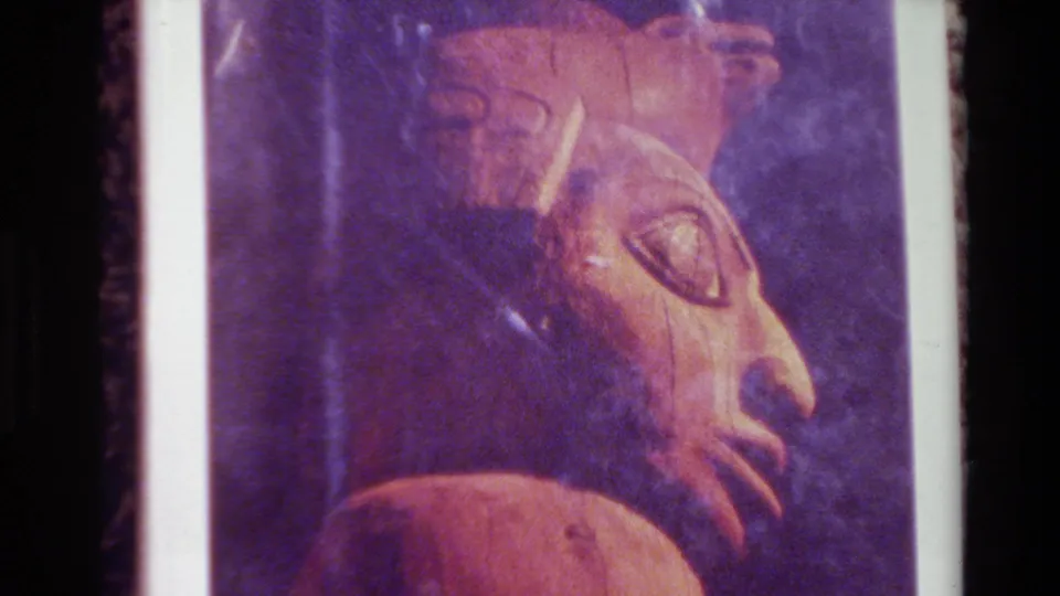 1980 Big Sur home-movie frame showing the lower portion of a brown carved face on a printed page, with dark background and film-edge artifacts.