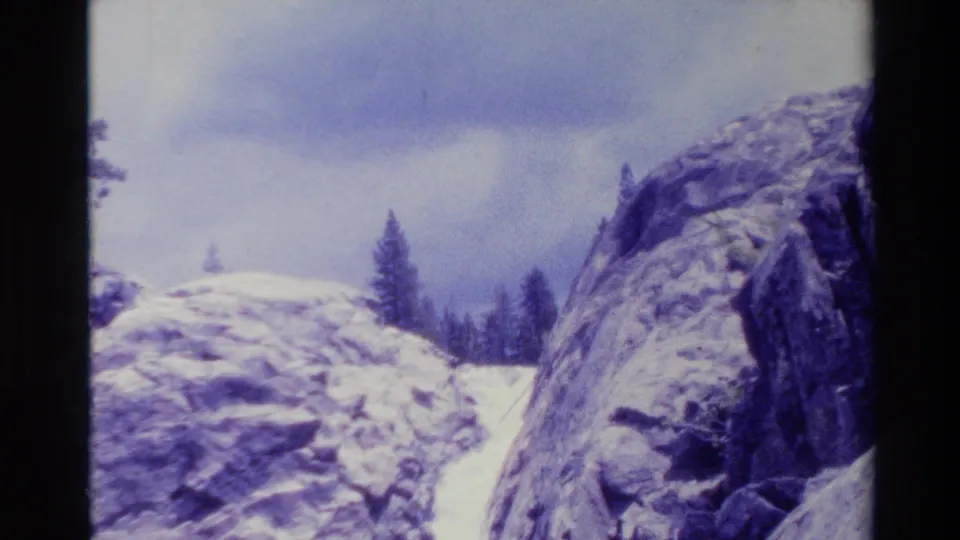 A rocky Big Sur landscape appears with dark pines, cloudy sky, pale ground, and a narrow waterfall-like streak in 1980 8mm footage.