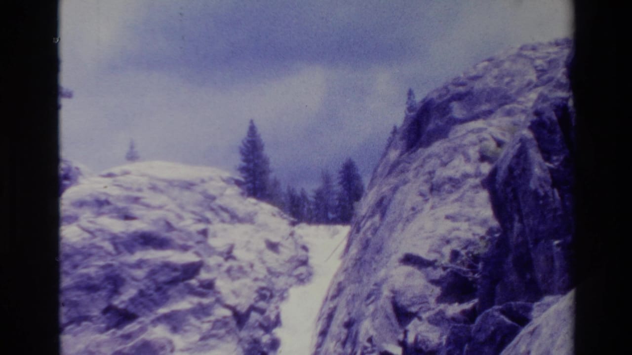 1980 Big Sur home-movie frame showing a narrow waterfall between snow-dusted rock formations with pine trees under an overcast sky.