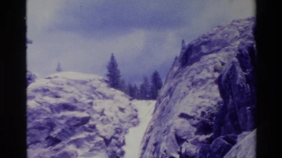 1980 Big Sur home-movie frame showing a narrow waterfall between snow-dusted rock formations with pine trees under an overcast sky.