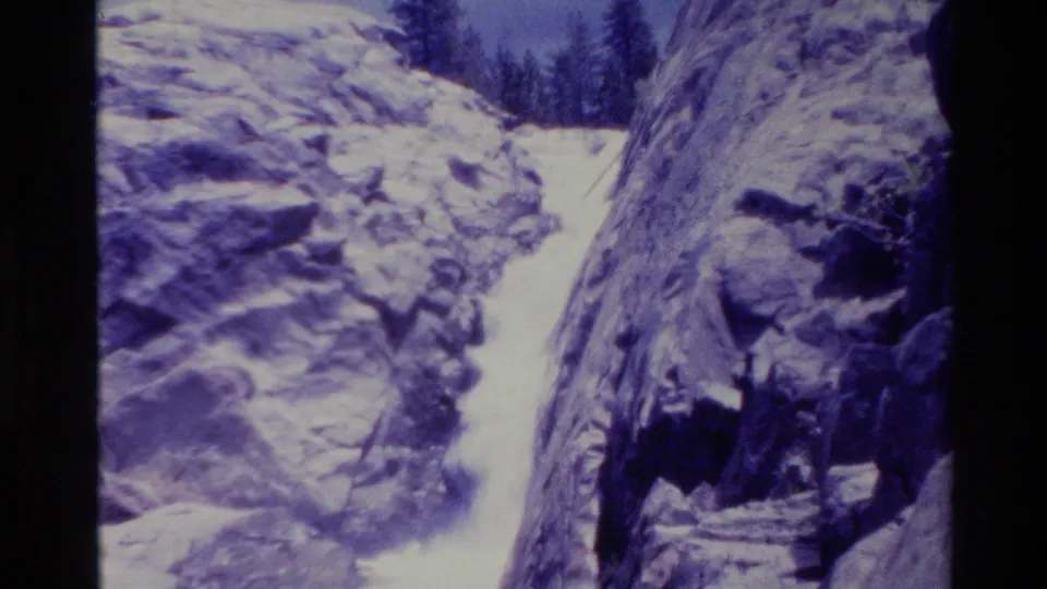 1980 8mm Big Sur frame with water flowing through a narrow rocky gap, snow-dusted walls, and trees along the canyon rim.