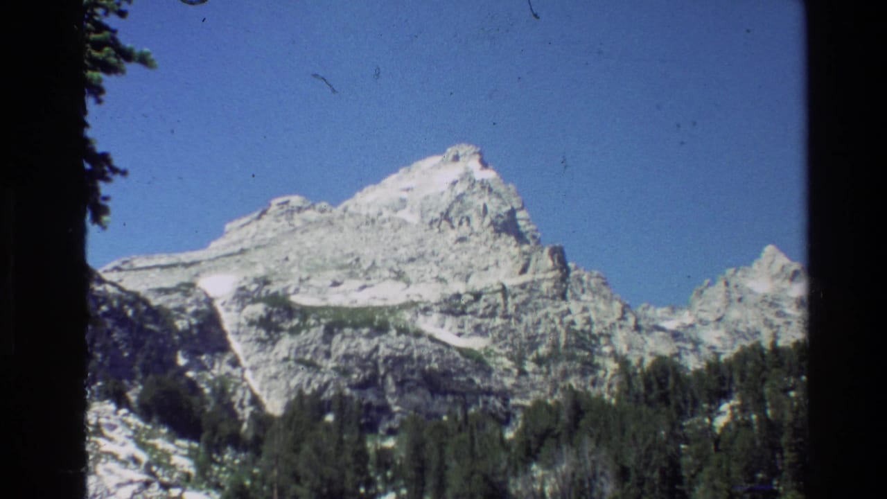 A Grand Teton mountain view shows pale rock, snow patches, dark evergreen forest, and clear blue sky in 1980 8mm footage.