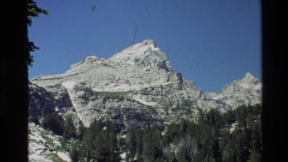 The 1980 Grand Teton home-movie frame holds on a snow-patched rocky peak with evergreen forest below and visible 8mm frame edges.