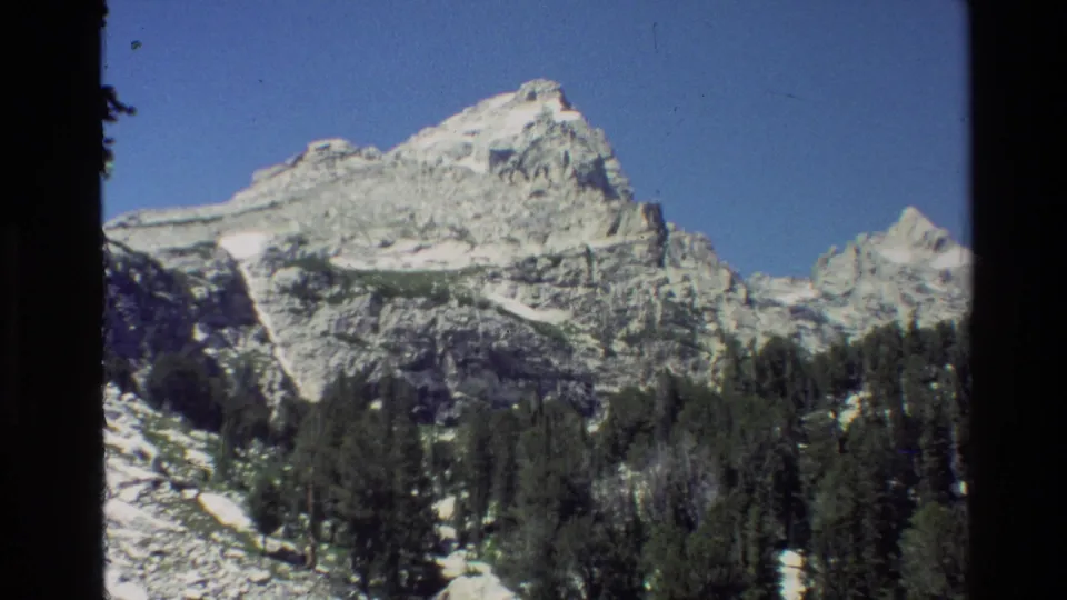 Grand Teton scenery fills the 8mm frame, with pale rock faces, lingering snow, evergreen trees, blue sky, and dark rounded borders.