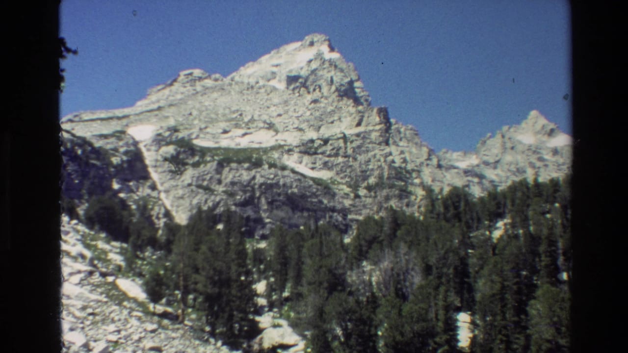 Rugged mountain peak with snow patches above pine trees and rocky slope.