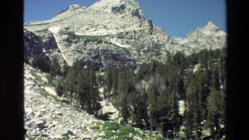 1980 Grand Teton 8mm frame showing rocky alpine terrain, dense pines, green grass, and snow below a pale peak.