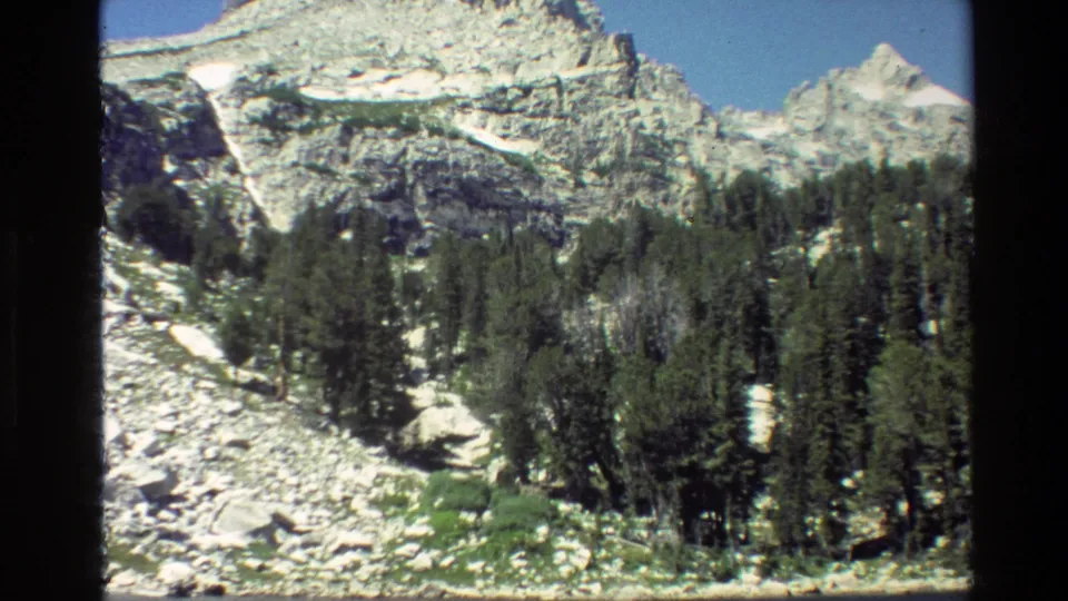 Final 1980 Grand Teton frame holding on a rocky mountain slope, conifer stand, snow patches, grass, and blue sky.