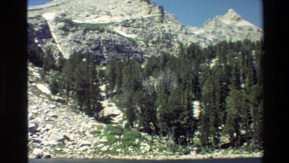 1980 8mm view of Grand Teton scenery with pine trees, pale rocks, snow patches, and distant mountains under a clear sky.