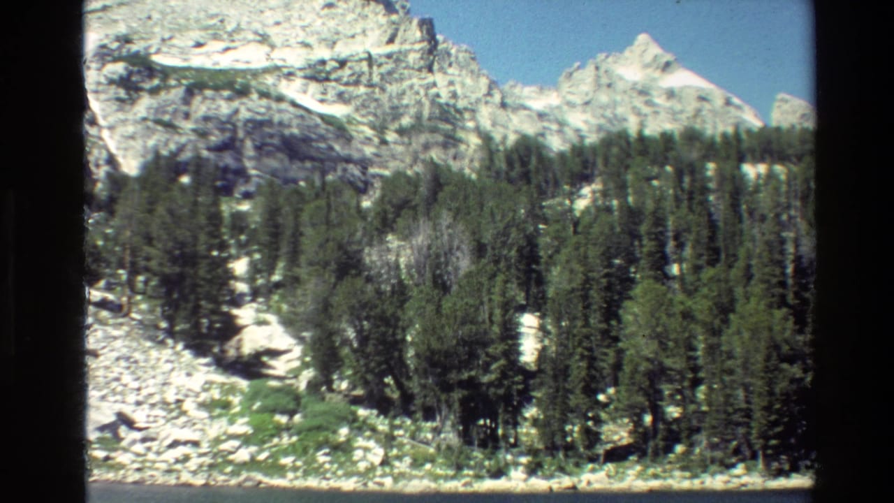 Grand Teton home-movie frame showing dense evergreen trees along pale rocks with snow-capped mountains rising behind them.