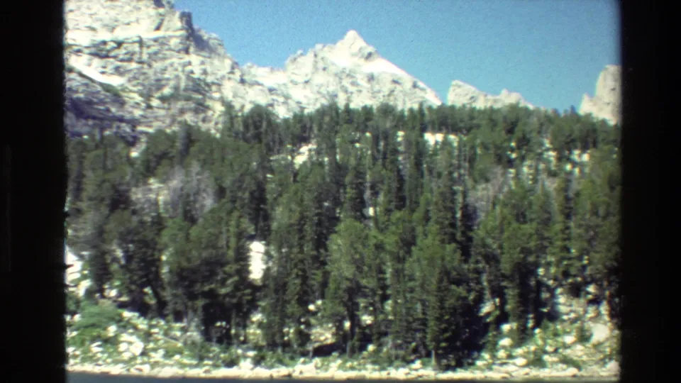 Vintage 8mm Grand Teton view of pine trees at a rocky lakeshore with bright snow-capped mountains and blue sky beyond.