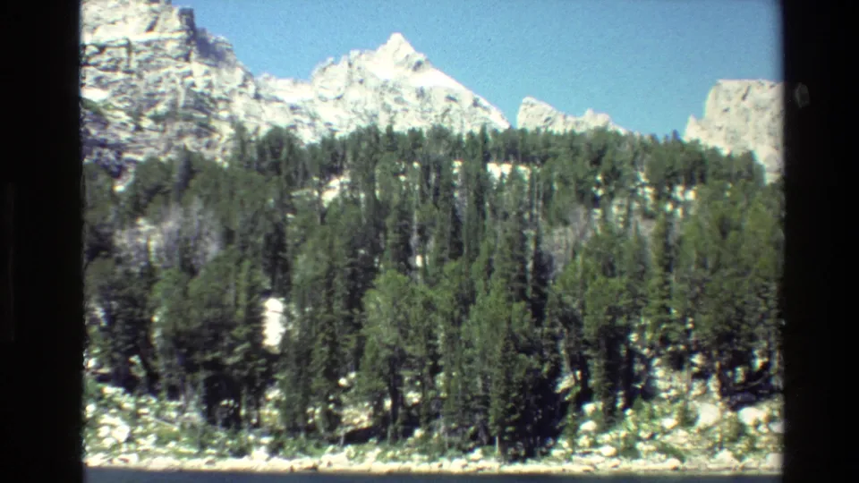 Grand Teton, Wyoming 8mm frame showing snowy peaks behind a dark evergreen forest and rocky foreground in bright sun.