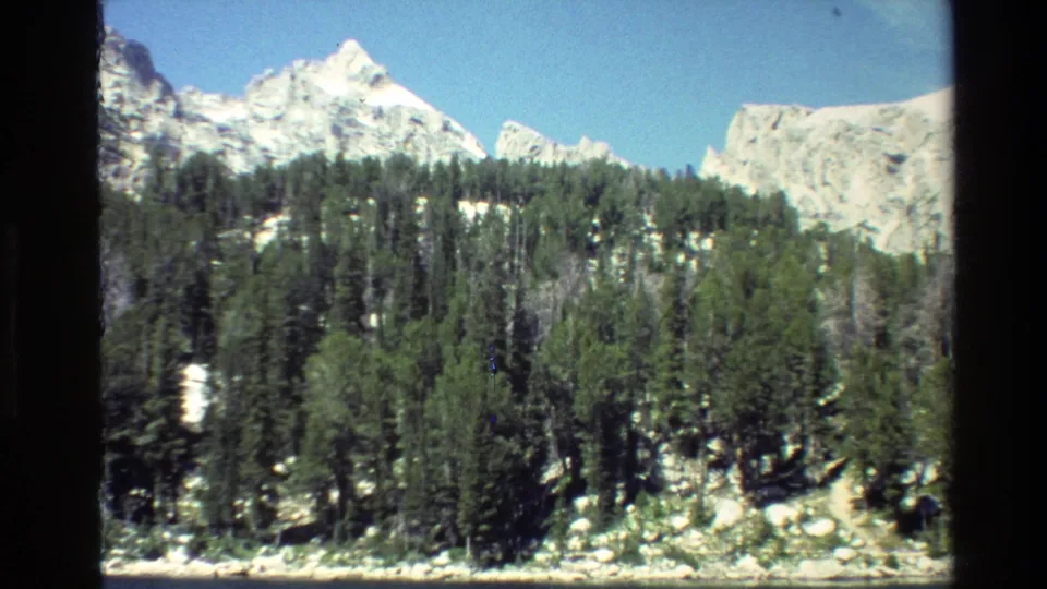 1980 Grand Teton home-movie frame with a lower forested ridge, rocky edge, and pale snowy mountains beyond.
