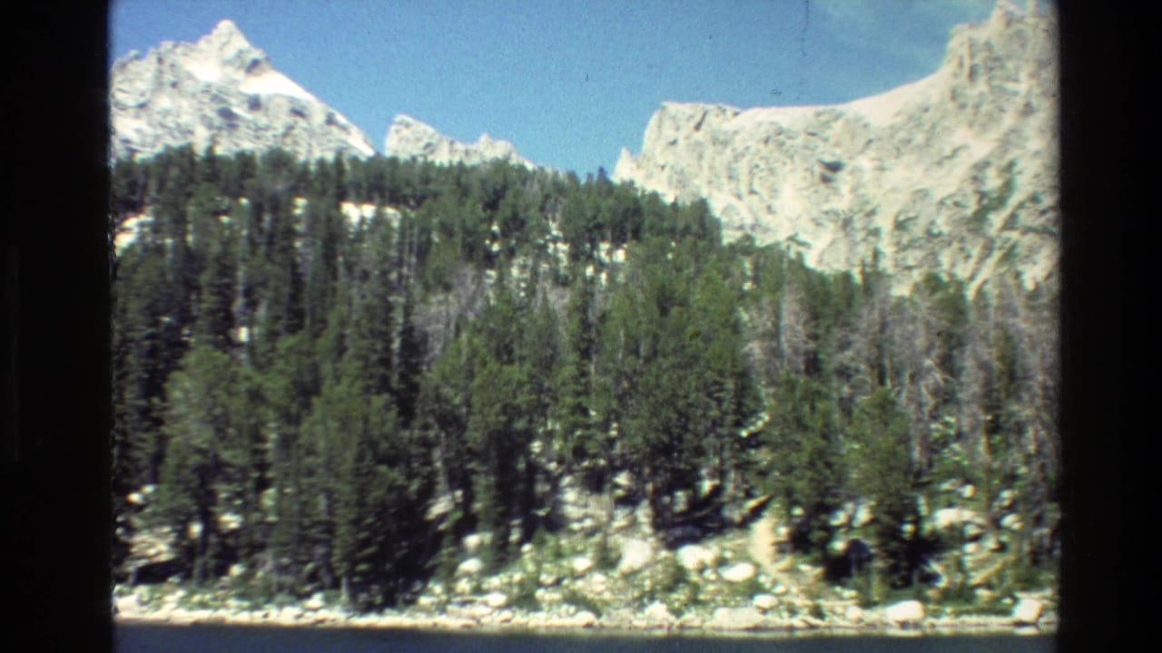 1980 8mm Grand Teton view with snow-capped peaks, dark pine forest, pale rocks, and a narrow waterline under clear blue sky.