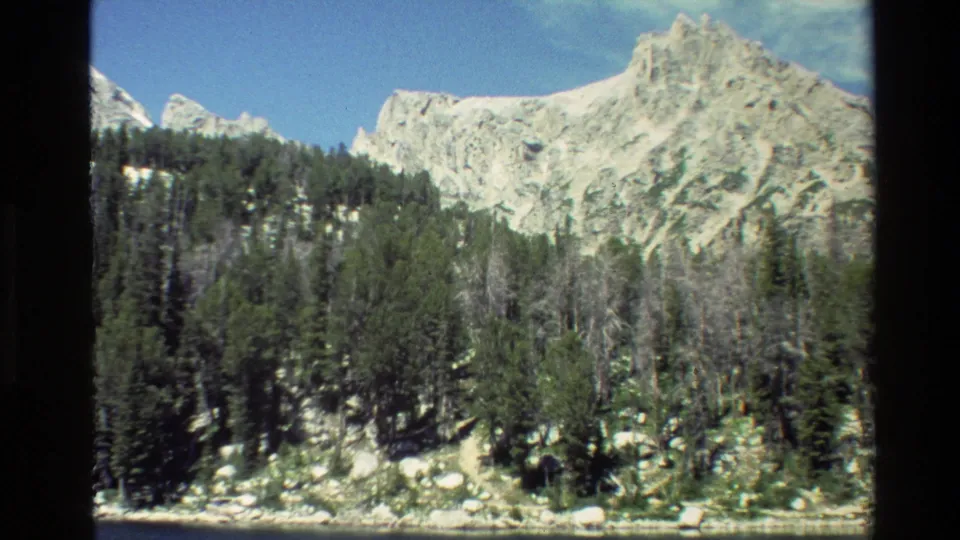 1980 home-movie frame of a rocky Grand Teton peak above green forest, boulders, and a narrow lakeside edge.