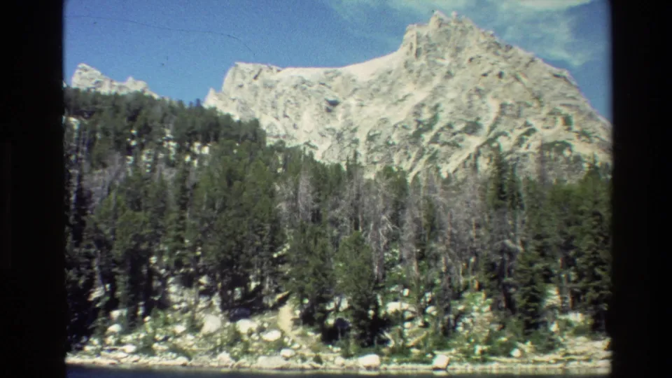 Grand Teton landscape frame showing rugged pale rock, evergreen forest, scattered boulders, and a small lakeshore band.