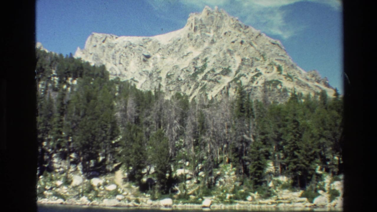 Grand Teton landscape frame with pale rocky summit, snow patches, evergreen trees, and a narrow strip of water at the base.