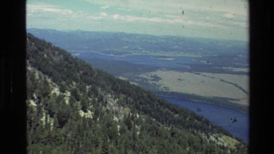 Grand Teton area overlook view on 8mm film, looking past a forested slope toward water, wetlands, valley floor, and distant mountains.