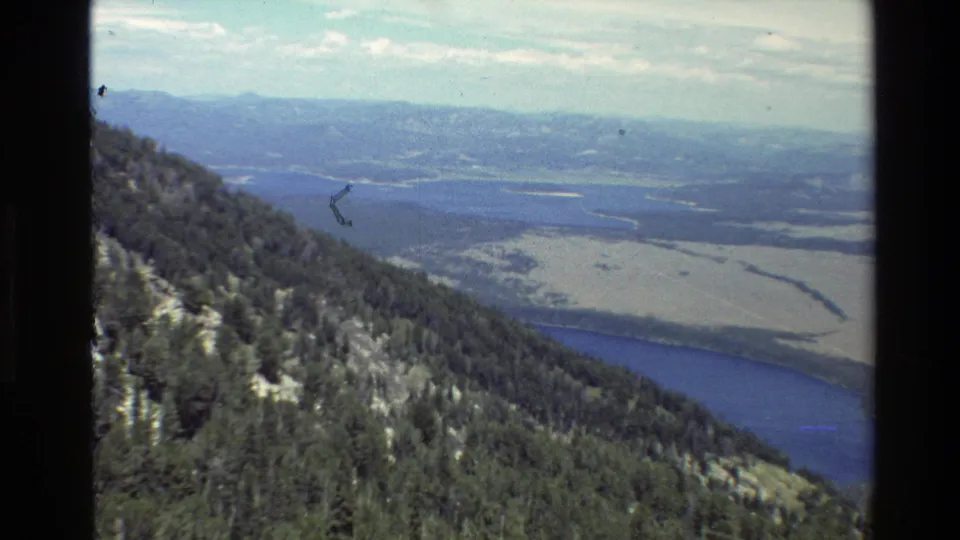 1980 Grand Teton 8mm frame from a high viewpoint, with evergreen hillside, blue water, open valley wetlands, and distant ridgelines.