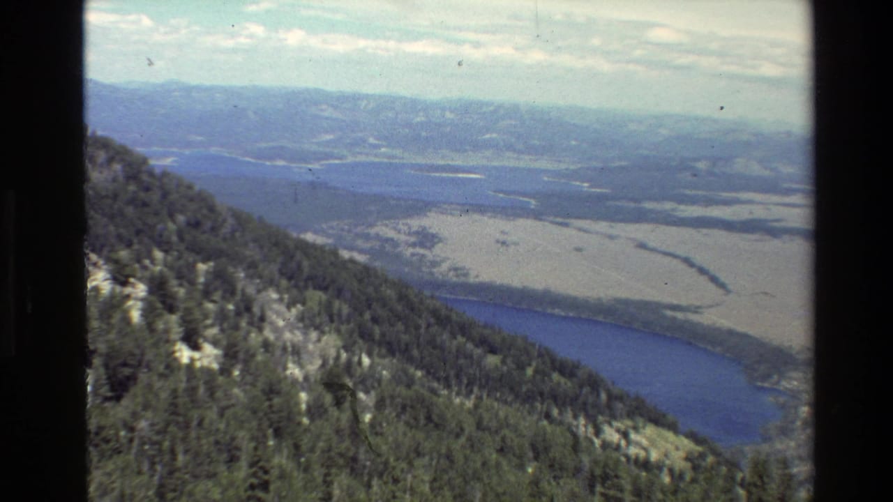 1980 Grand Teton, Wyoming view with dark conifer forest on a sloping mountainside above lake water and distant valley terrain.