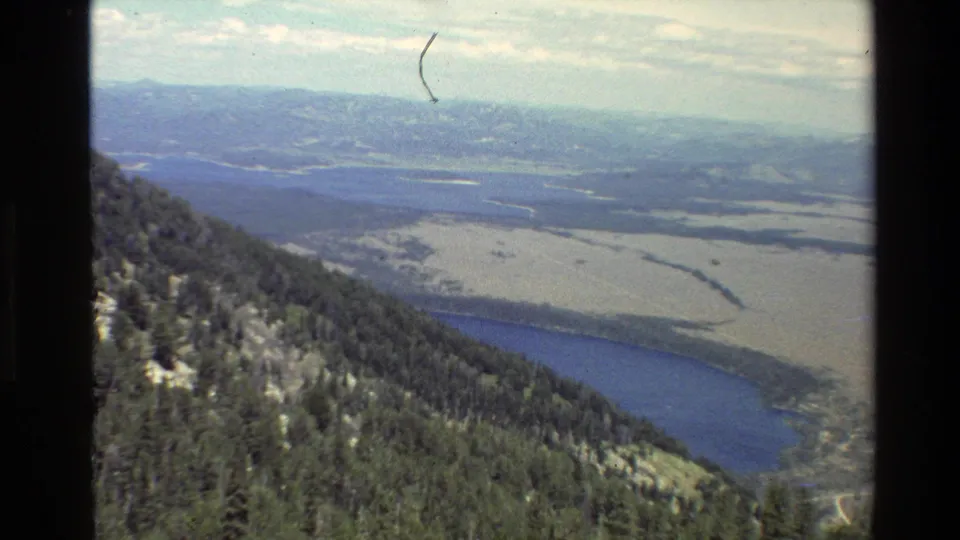 1980 Grand Teton, Wyoming frame with conifer slopes at left, lake shoreline below and a broad valley stretching toward distant hills.