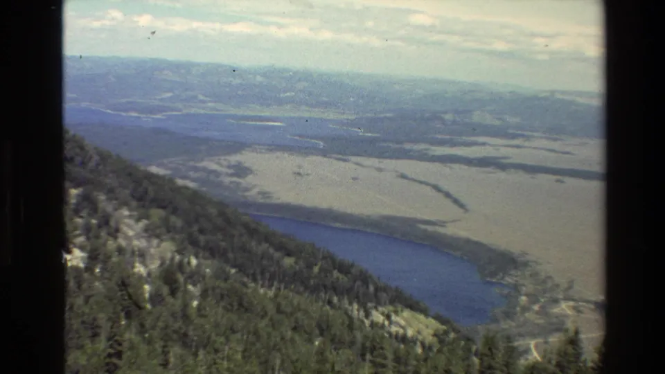 1980 Grand Teton, Wyoming 8mm view of forested mountain terrain, blue lake water, pale valley plains and distant ridgelines.