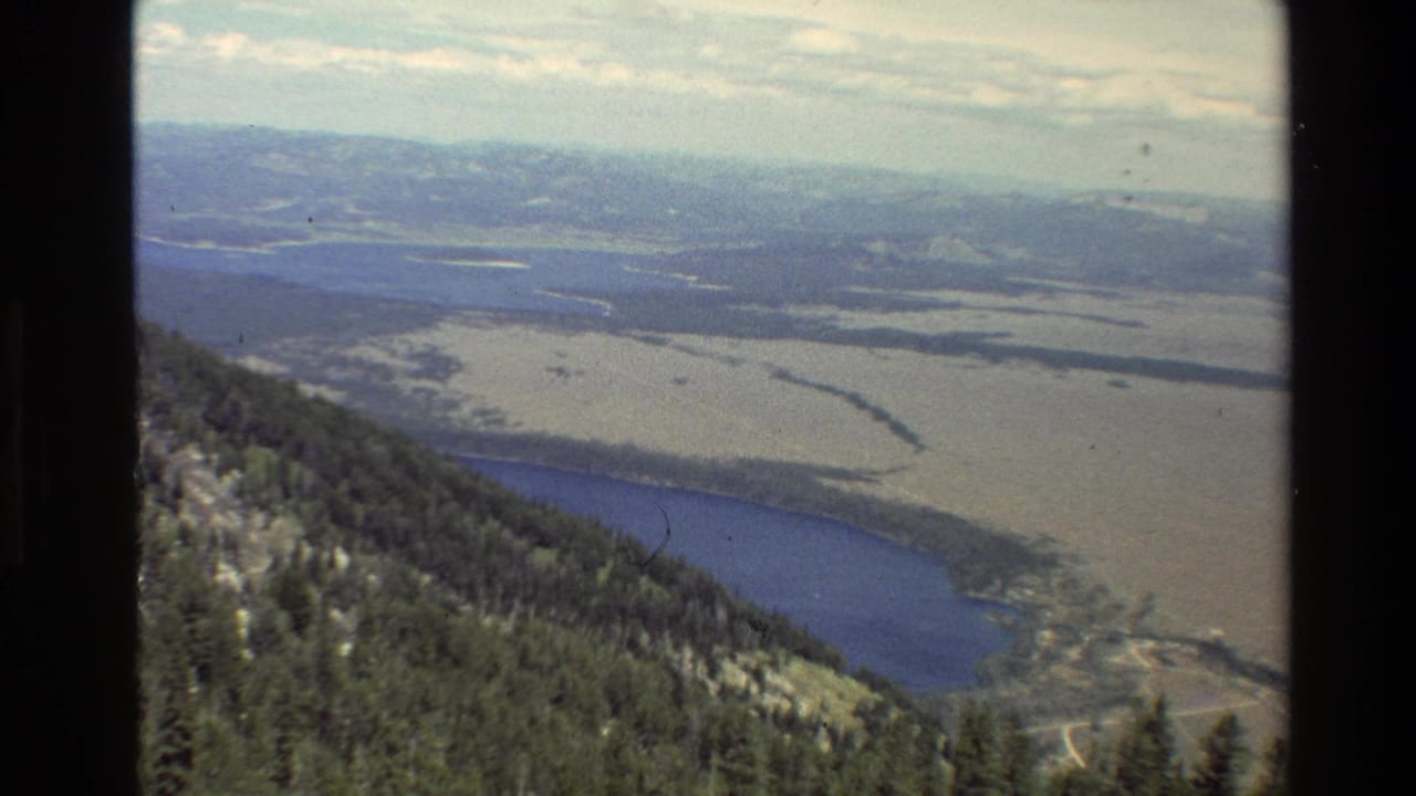 1980 Grand Teton home-movie frame showing a forested slope above a blue lake, open valley flats, and hazy mountain ridges.