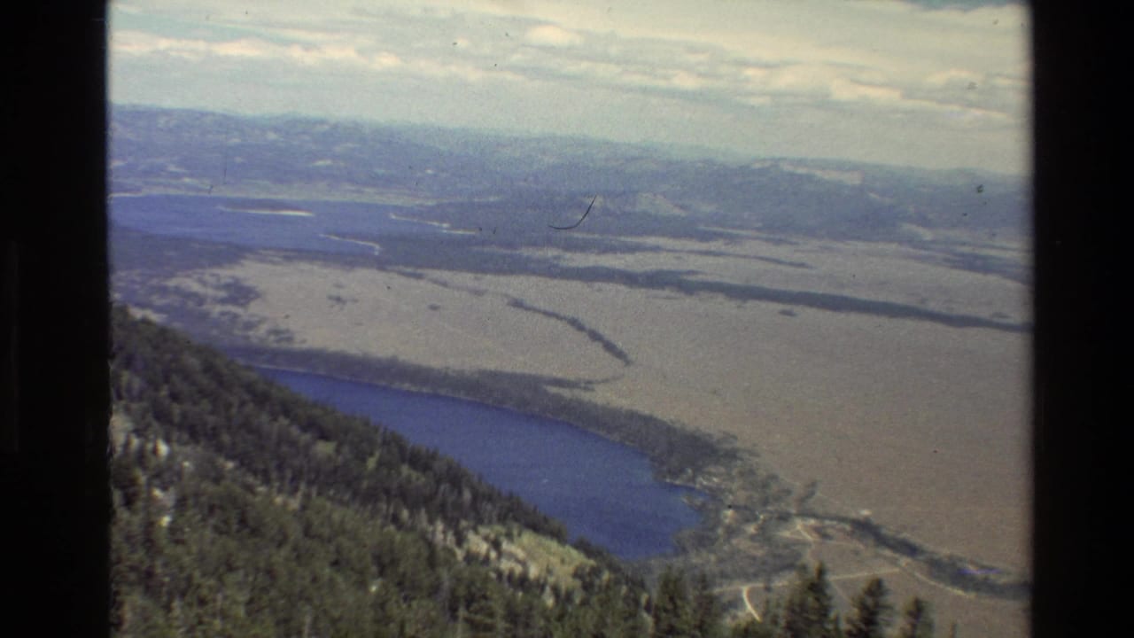 High view of a wooded slope, blue lake, pale valley floor, river channels, and distant mountains.