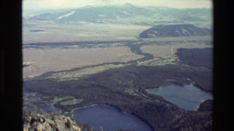 Grand Teton region frame showing dense forest around blue lakes, a winding river across the valley, and soft distant ridgelines.