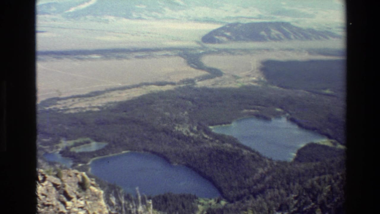 1980 Grand Teton area home-movie frame showing two blue lakes below a forested slope, with valley flats and distant ridges beyond.