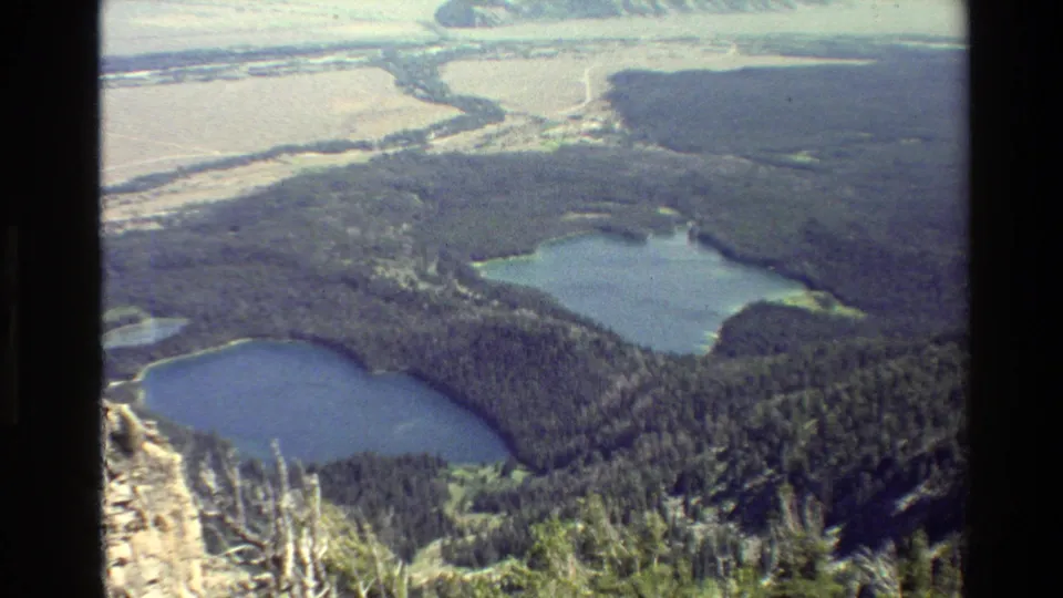 Grand Teton, Wyoming 1980 home-movie frame with two lakes below a wooded slope and pale valley terrain in the background.