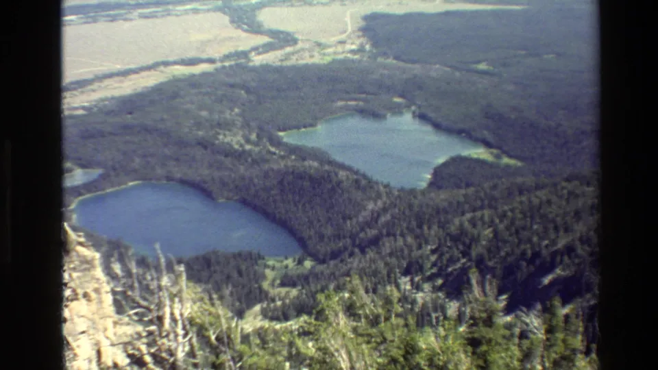1980 Grand Teton area frame showing blue lakes and evergreen forest from a high overlook, with bright valley flats in the distance.
