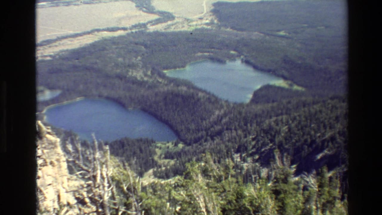 1980 Grand Teton home-movie frame showing two blue alpine lakes below evergreen forest and rocky slopes from a high overlook.