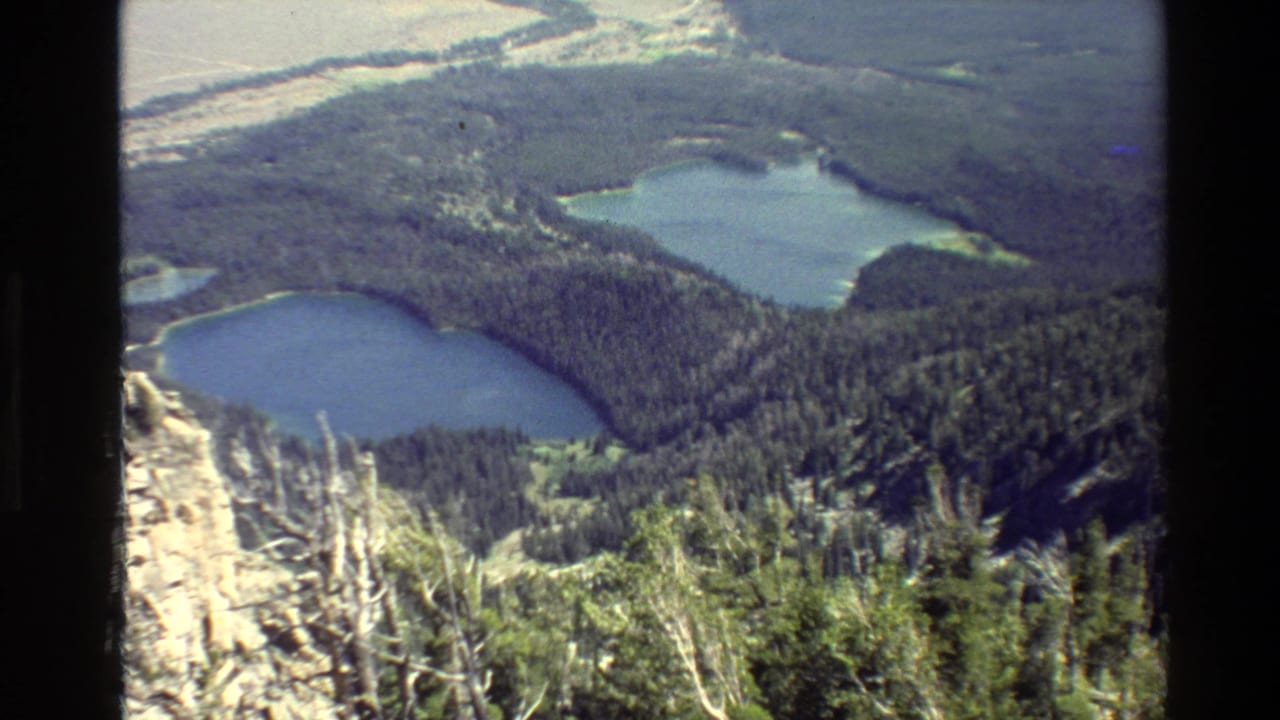 1980 Grand Teton footage frame with blue alpine lakes, dense conifer forest, and bright rocky ground viewed from above.