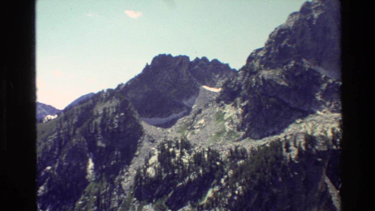 1980 8mm Grand Teton view of rugged rocky peaks, pale sky, lingering snow, and forested lower slopes.