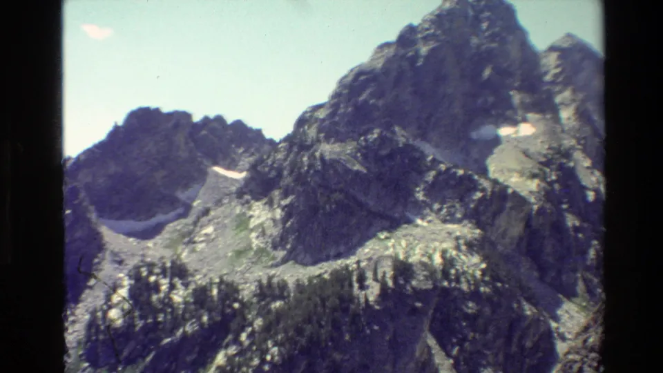 1980 Grand Teton home-movie frame showing a craggy summit, darker forest below, and lingering snow across rocky terrain.