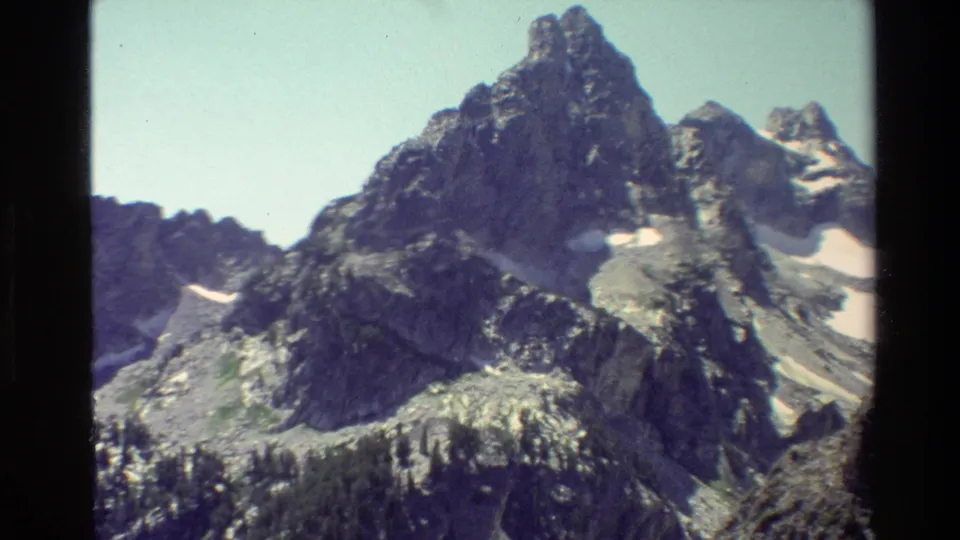 Grand Teton landscape frame with a prominent rocky peak, adjacent snow patches, evergreen forest, and visible 8mm edge darkening.