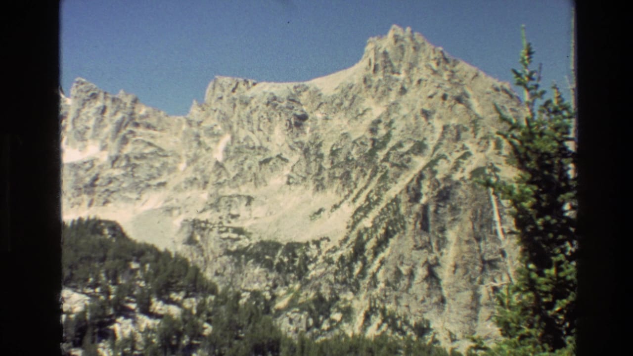 1980 8mm view of Grand Teton peaks in Wyoming, with snow on rugged rock faces and evergreens along the lower frame.