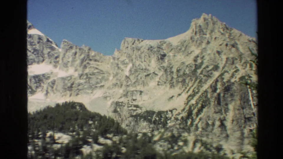 1980 8mm home-movie frame of the Grand Teton range, with snow patches on rocky peaks and evergreen-covered slopes below.