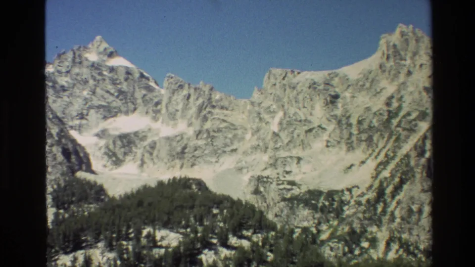 Grand Teton landscape in 1980 8mm footage, showing multiple snow-dusted peaks above evergreen slopes and a clear sky.