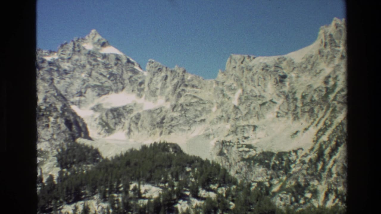 1980 Grand Teton 8mm frame showing rocky peaks, lingering snow, and forested valley slopes beneath a clear blue sky.