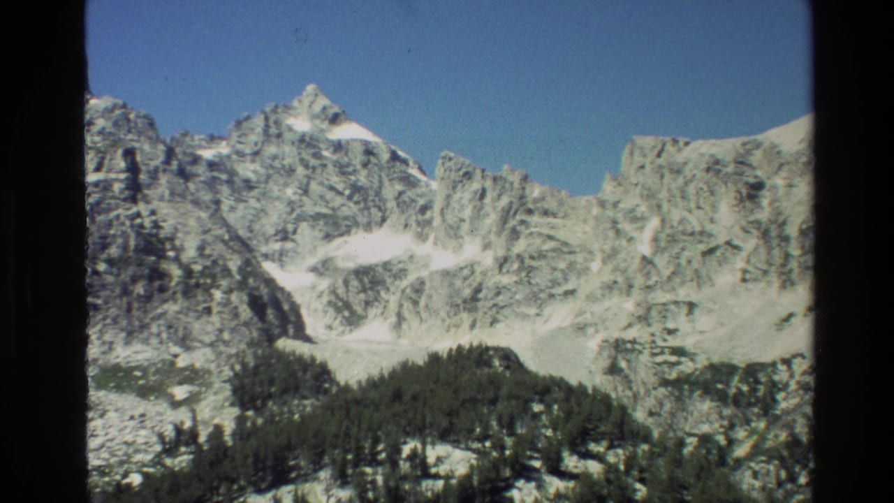 Grand Teton, Wyoming landscape frame with rugged stone ridges, white snowfields, and dark conifers below.