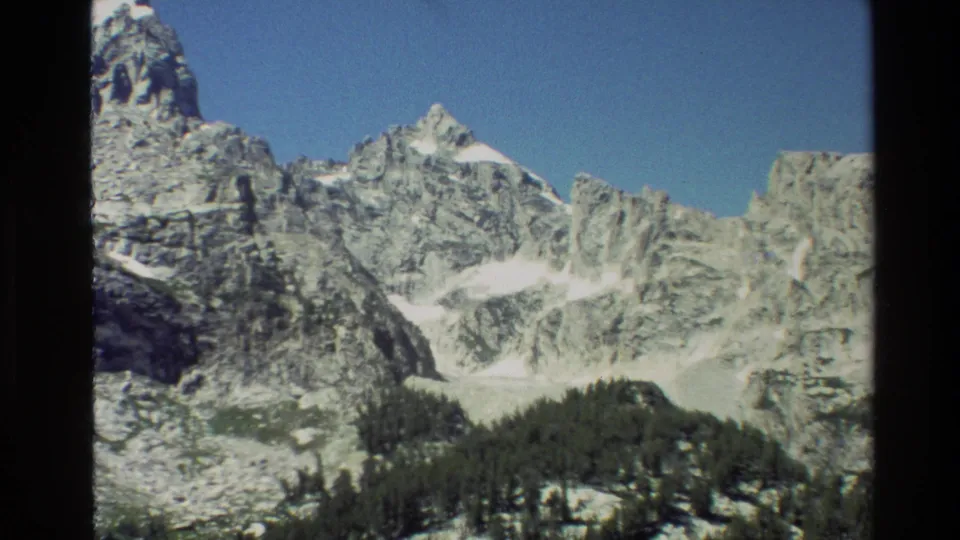 Visitor-shot 8mm view of Grand Teton peaks with pale snowfields, dense trees, and rugged exposed rock.