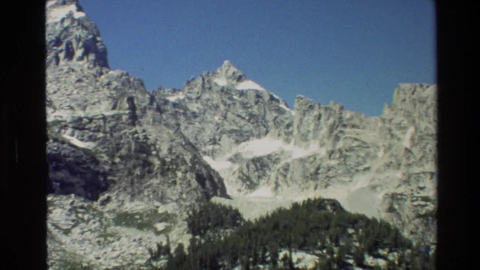 Grand Teton landscape frame showing snow-streaked peaks, rugged cliffs, and a shadowed evergreen ridge.