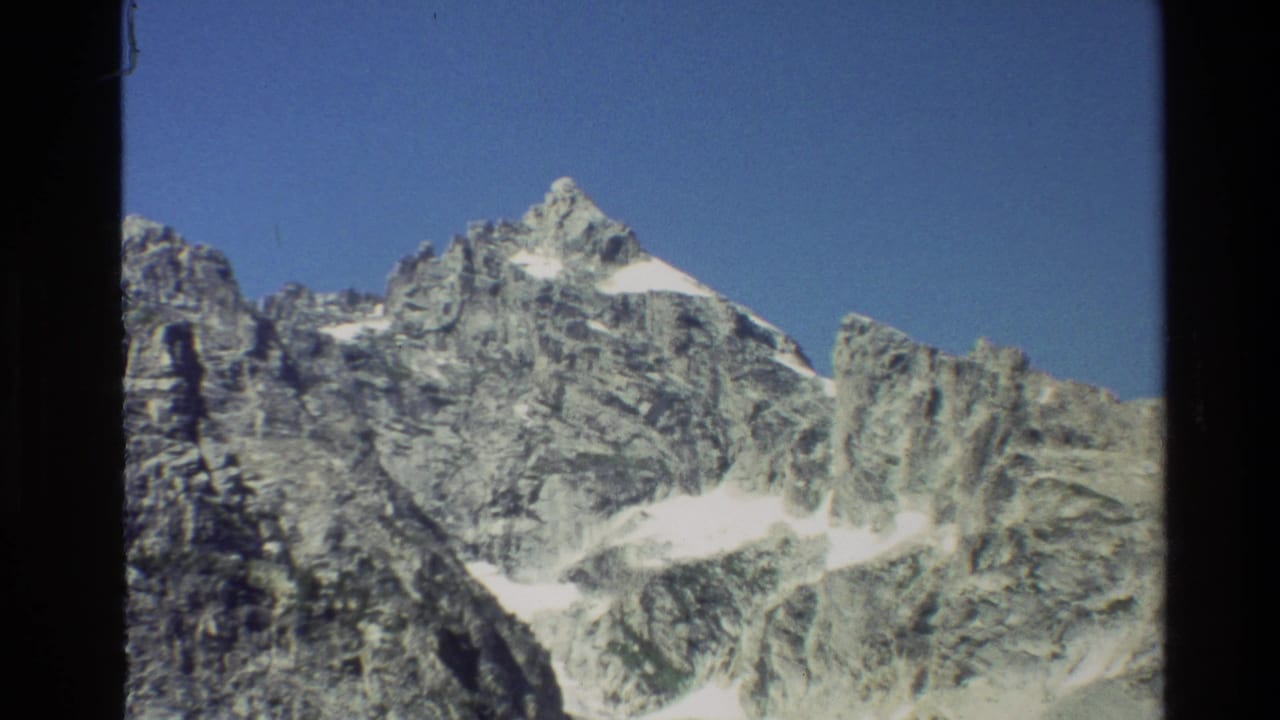 1980 8mm view of the Grand Teton range, showing rocky snow-streaked peaks beneath a clear blue sky.