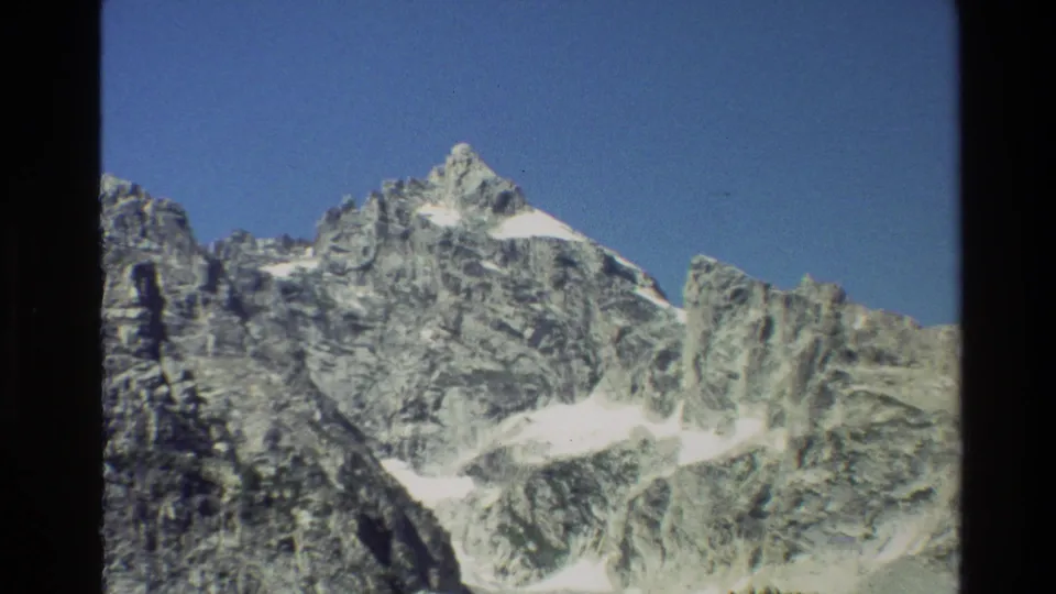Grand Teton mountain ridge in 1980 home-movie footage, framed by dark 8mm borders and a clear blue sky.