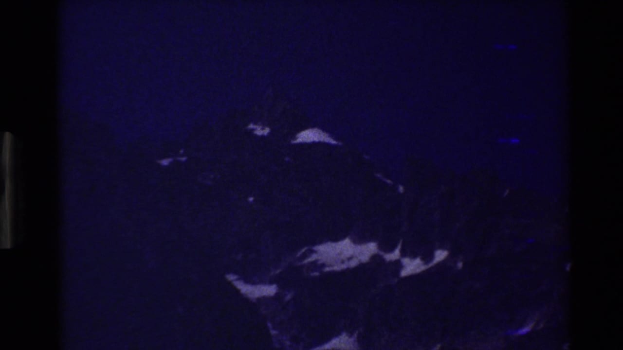 Underexposed 1980 8mm frame of Grand Teton with a blue cast, dark gate edges, rocky peak, and scattered snow patches.