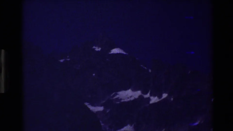 Underexposed 1980 8mm frame of Grand Teton with a blue cast, dark gate edges, rocky peak, and scattered snow patches.
