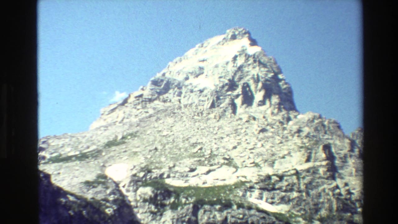 1980 8mm frame of Grand Teton showing a rugged sunlit summit, scattered snowfields, and clear blue sky within visible film edges.