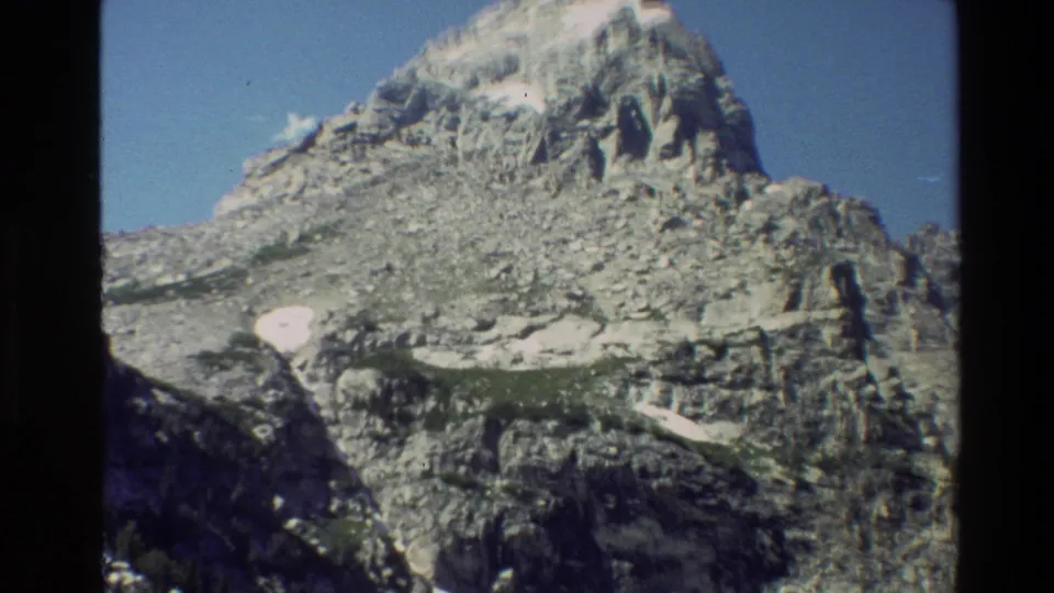 1980 8mm view of Grand Teton's rocky face with visible snowfields, blue sky, and the dark edge of the scanned film frame.